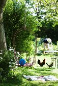 Idyllic garden - wire chair hanging from tree on rope, hens beneath bistro table and person gardening in background