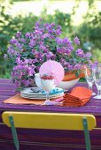 Vorbereitungsstadium eines sommerlich gedeckten Gartentisches mit Bougainvillea auf violettem Tischtuch und Stuhllehne in der Komplementärfarbe Gelb
