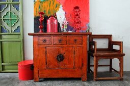 Ornaments on old Oriental cabinet in front of modern artwork and next to wooden chair in simple room