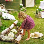 Von Baumstämmen begrenztes Gemüsebeet mit Blumenkohl und Salat; kleines, barfüssiges Mädchen bei der Beetpflege