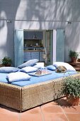 Wicker couch with blue cover and cushions on sunny terrace; view into kitchen of modern, Mediterranean house in background