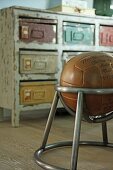 Leather ball in metal chair frame in front of vintage chest of drawers with metal boxes in place of drawers