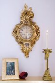 Lit candle in brass candlestick on surface and gilt-framed wall clock on pastel wall