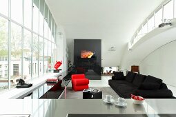 View across stainless steel kitchen counter to open-plan interior with black sofa and red designer armchair next to huge window