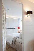 Lit sconce lamp in hall next to open, white louver door and view into modern bedroom
