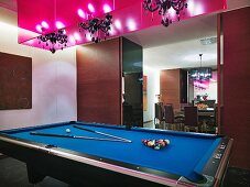 Pool table with cornflower blue felt set up for game below chandeliers in hot pink light boxes