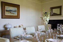 Simple yet festively set table and chairs with pale, checked upholstery in traditional dining room with fireplace and landscape painting above sideboard.