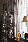 Wire tailor's dummy hung with innumerable necklaces next to antique dressing table with perfume bottles in front of floral curtains at window