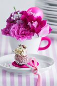 A chocolate on a teaspoon on a saucer, in front of a cup holding pink flowers and a name badge