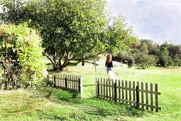 Green, summer landscape with picket fence and woman walking across field beyond