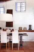 Dining area with massive table and upholstered white chairs in front of collection of jugs and stoneware pots on shelf on whitewashed wall with lattice windows in gable end wall