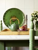 Tea set on tray in front of green, painted metal platter leaning on white tiled wall