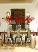 Retro metal chairs around wooden table with branches of berries in vases in front of tapestry hanging on wall