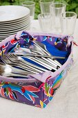 Patterned plastic container of cutlery in front of stacked plates and glasses on tablecloth in garden
