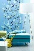 Polsterhocker in Aquafarben vor Designer-Stehleuchte, im Hintergrund blauer Wandbehang mit Magnolienblüten-Motiv