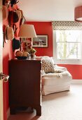 View through open door into red-painted room with chest of drawers and white armchair below window