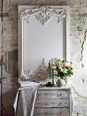 White, decorative, carved panel on wall behind bouquet of roses and bolt of embroidered fabric on chest of drawers delicately painted in soft colours