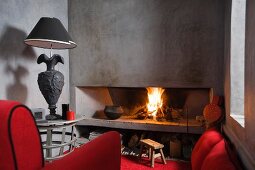 Brennendes Kaminfeuer in grauem Raum mit roten Polstermöbeln und schwarzer Tischlampe mit Fuss in Form einer Amphore