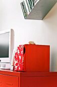 Rotlackiertes Sideboard mit weißem Flachbildschirm und floralgemusterter Box; darüber schräges Metallwandboard mit DVDs