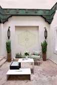 Oriental courtyard with white designer table on terracotta floor, geometric painting in niche, symmetrical sconces, vase-shaped planters and wooden panels painted dark green running around upper wall