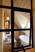 Blick durch ein altes Eisenfenster in das elegant-gemütliche Badezimmer eines Landhauses mit holzvertäfelter Dachschräge