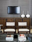 White vases on brown and black sideboard and flat-screen TV on grey wallpapered wall; two elegant armchairs with woven backs in foreground