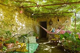 Idyllische Gartenterrasse mit Weinpergola und Hängematte mit darin lesendem Jungen; davor ein Tagesbett mit verspieltem Metallgestell