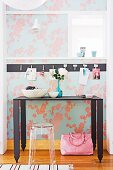 Black, metal console table and transparent, plastic stool against floral wallpaper