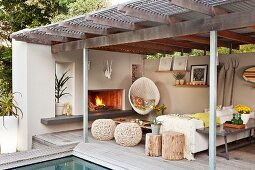 Blick auf Sitzecke auf überdachte Terrasse am Pool mit Kaminfeuer, Hängesessel & Hockern aus Naturmaterialien