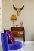 Purple velvet chair with retro-style cushion in front of Baroque bureau, table lamp and animal skull