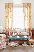 Comfortable, romantic living room with floor-length floral curtains at bright window behind red and white striped sofa with many scatter cushions and light blue ottoman