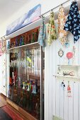 Sammlung farbiger Glasflaschen in Raumteilerregal zwischen aufgehängten, gemusterten Tüchern und Kitschgegenständen