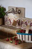 Chip wood basket of strawberries on side of stone sink; water running from wall-mounted taps above old, patterned wall tiles