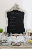Chalkboard in ornamental frame behind collection of crystal vessels on rustic console table