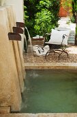 Natural stein water feature with three spouts in a Mediterranean garden; patio chairs on gravel in the background