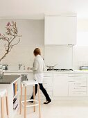 Woman walking in front of purist, white kitchen counter; bar stools at island block