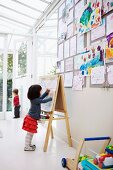 Little girl drawing on easel and collection of child's drawings pinned on wall in white interior with glass ceiling