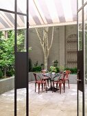 Blick durch offene Terrassentür auf farbige Metallstühle mit Retro-Flair an rundem Tisch im Innenhof