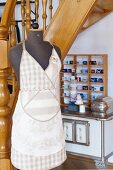 Tailor's dummy wearing linen apron in sales room; shelves of rolled, fabric ribbons in background