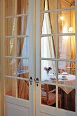 View of set breakfast table below window with gathered curtains through double interior doors with lattice windows