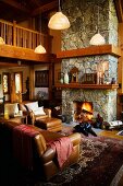 Gediegene, braune Ledersofagarnitur vor Kaminfeuer in offenem, rustikalem Wohnzimmer und Galerie aus Holz in warmem Braunton