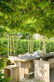 Rustic dining table and chairs made from wooden boards below green, vine-covered pergola