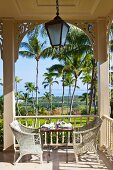 White rattan chairs at set table on colonial-style veranda with view into garden of palm trees