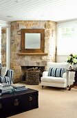 Rustic, rough stone wall with open fireplace and square mirror on chimney breast in living room with Hamptons-style furniture; books on vintage trunk in foreground