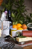 Stillleben mit Holztablett neben Rotweingläsern und -flaschen, Käse, Wurst und Senfglas; im Hintergrund eine Obstschale mit Orangen und Apfel