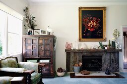 Armchair next to colonial-style cupboard and rustic fireplace below framed picture in living room