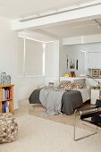 Doppelbett mit Textilien in Grautönen vor halbhoher Trennwand, Wohnbereich mit Bücheregal und Klassikerstuhl im Vordergrund