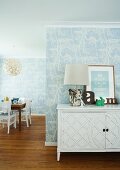 Table lamps with lampshades in natural colors on a white sideboard with carved door in front of a wall with bright blue wall paper with a floral pattern and a view into an open dining room