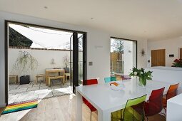 Colourful plastic chairs around white dining room in front of open terrace doors with view of courtyard