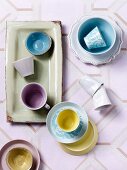Blick auf pastellfarbene Tassen, teilweise auf Schalen und Untertassen auf gemusterter Tischdecke im Retrostil